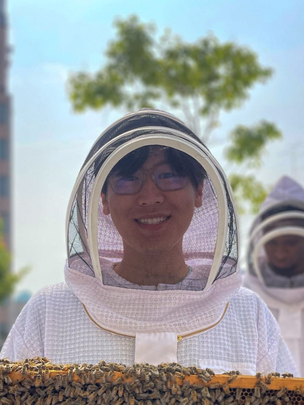 A person wearing a white beekeeper suit with protective mesh headgear, smiling while wearing glasses. Honeybees are visible on what appears to be a hive frame at the bottom of the image. Another beekeeper can be seen in the background. The image is overlaid with text reading 'Beekeeper enthusiast'