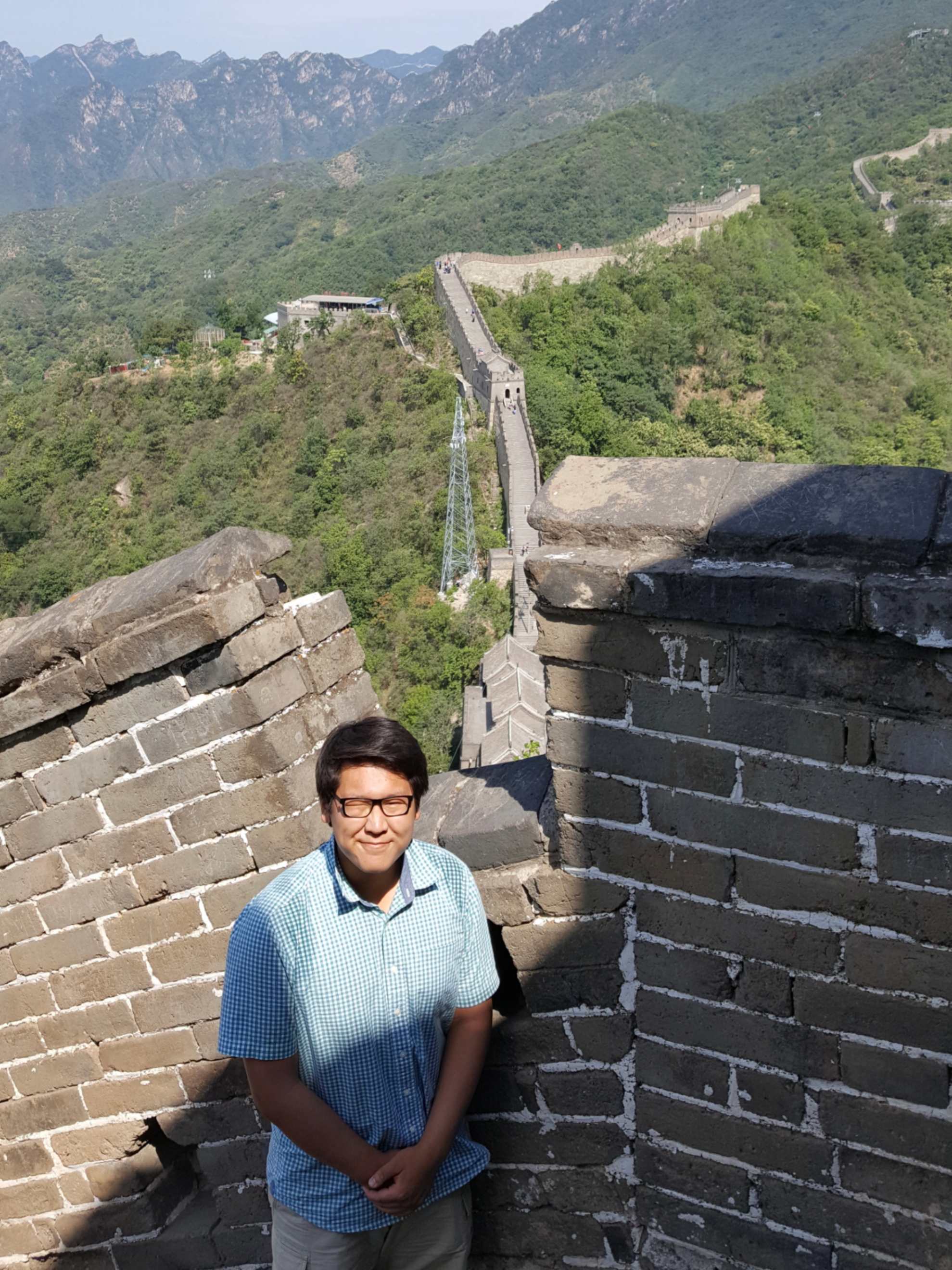 A person standing on the Great Wall of China wearing a light blue checkered short-sleeve shirt and glasses, smiling at the camera. The ancient stone walls extend into the distance through green mountainous terrain. The image is overlaid with text reading 'Purdue Study Abroad Summer 2017'