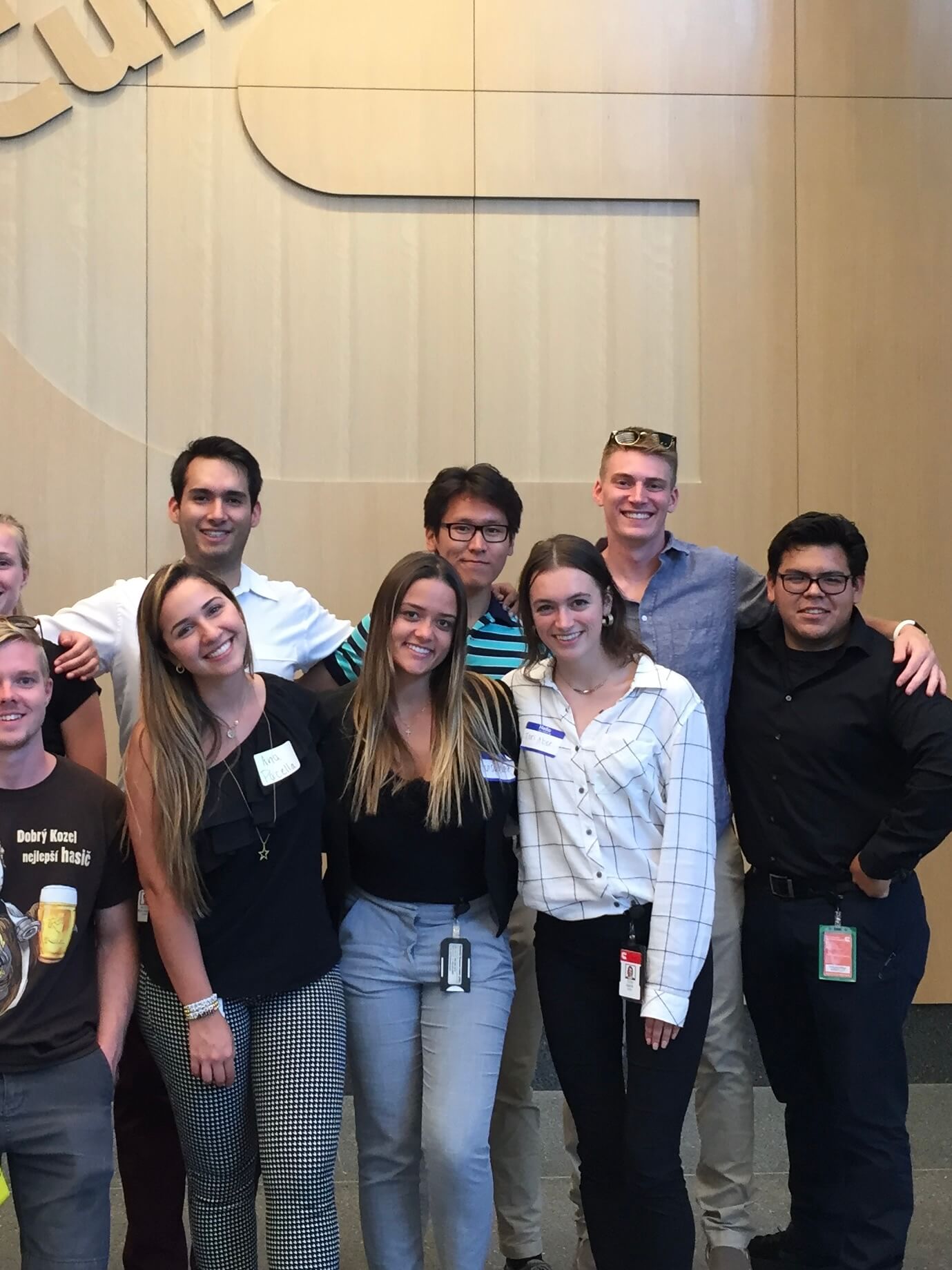 A group of Cummins interns standing together in an office hallway with beige walls. The group of eight people is dressed in casual business attire ranging from t-shirts to button-downs, with some wearing ID badges. Some are wearing glasses and sunglasses, and they are arranged in two informal rows. The image is overlaid with text reading 'Cummins Emissions Internship Summer 2019'