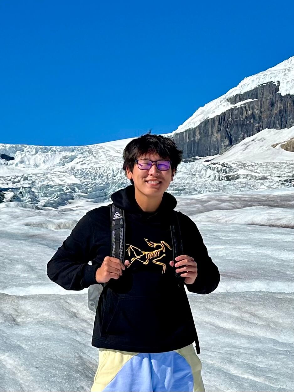 A person standing on a glacier wearing a black Arc'teryx hoodie and purple glasses, smiling at the camera. The backdrop features dramatic ice formations and snow-covered mountain cliffs. The image is overlaid with text reading 'A little about me'