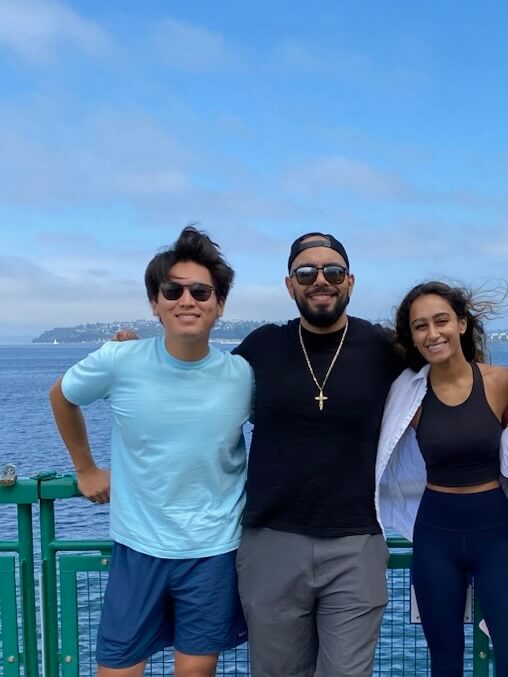 Three people standing together by a waterfront railing with a body of water in the background. The person on the left wears a light blue t-shirt and sunglasses, the person in the middle wears a black shirt and baseball cap with sunglasses, and the person on the right wears athletic wear. The image is overlaid with text reading 'Microsoft Finance Full-Time 2021 - Present'