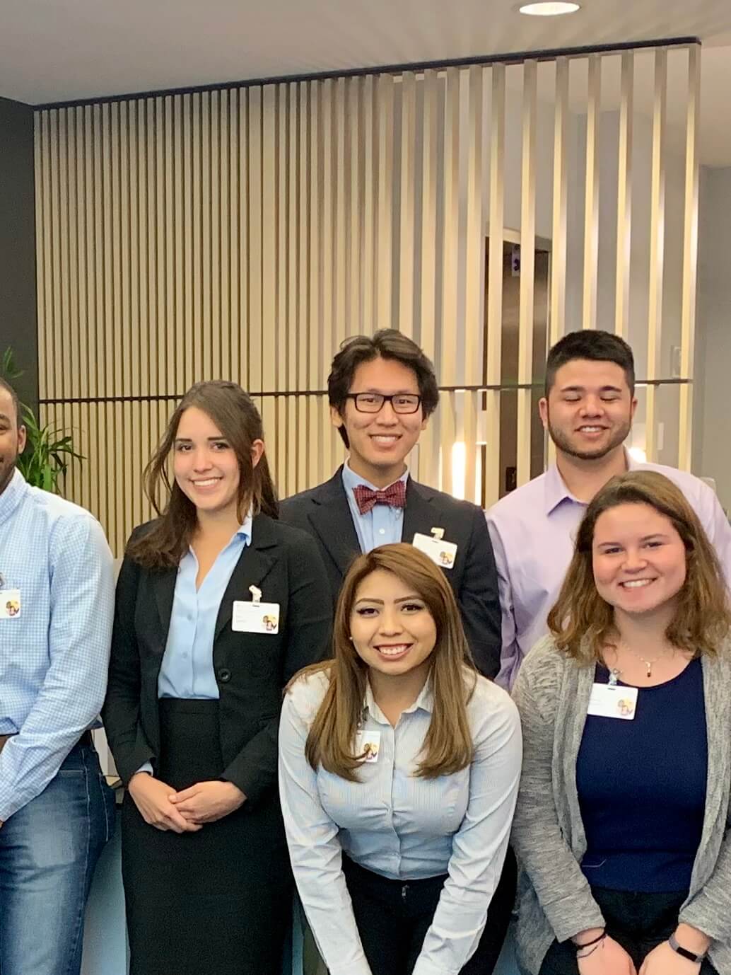 A group of Microsoft interns in business attire wearing ID badges, standing together in an office setting with wooden paneled walls. One person wears a suit with a bow tie, others wear button-down shirts and business casual attire. The image is overlaid with text reading 'Microsoft Finance Internship Summer 2020'