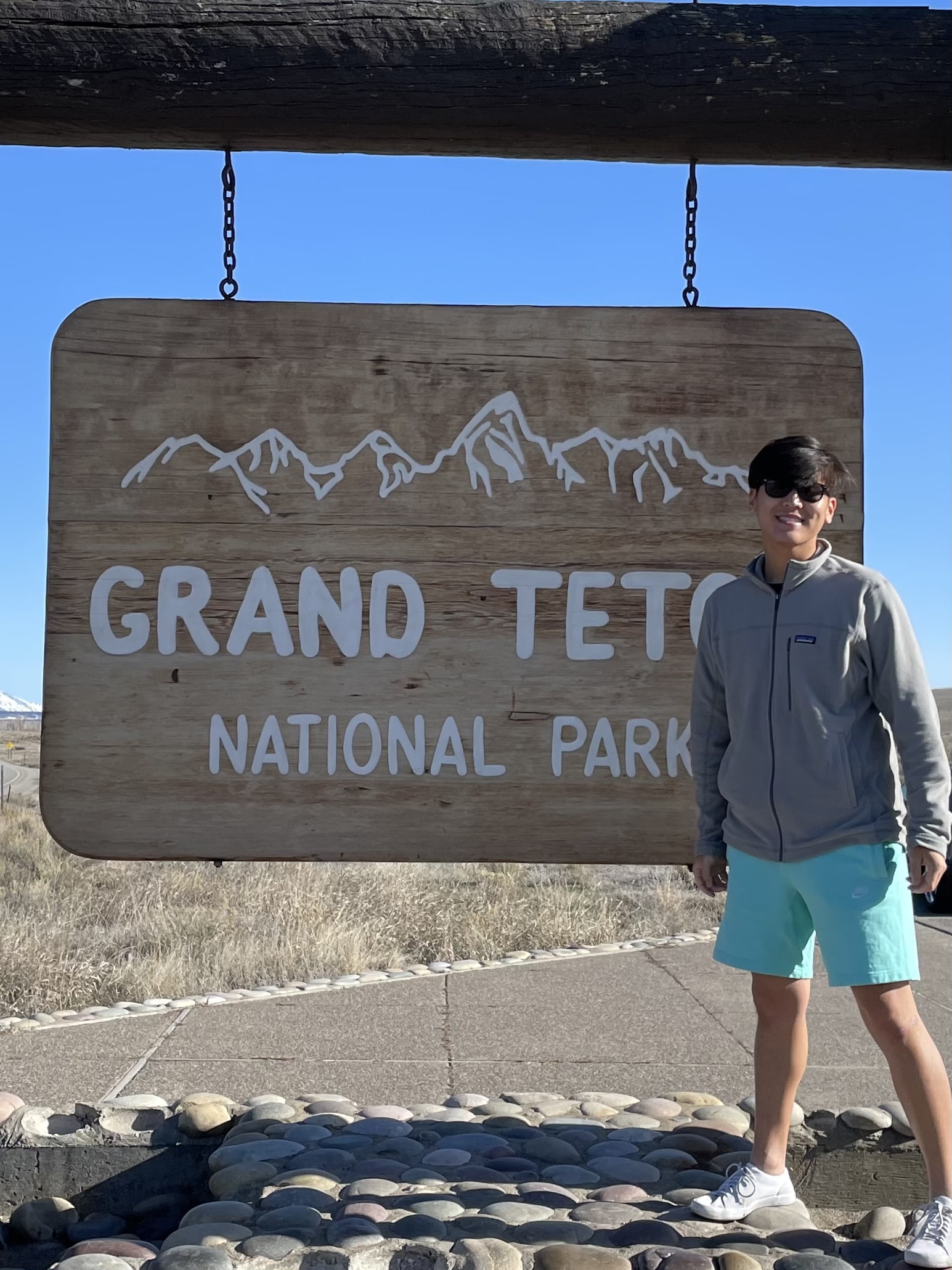 A person standing next to the Grand Teton National Park entrance sign wearing a gray fleece jacket, turquoise shorts, sunglasses, and a baseball cap. The wooden park sign features a mountain range design and is mounted on decorative stone work. The image is overlaid with text reading 'National Parks adventurer'