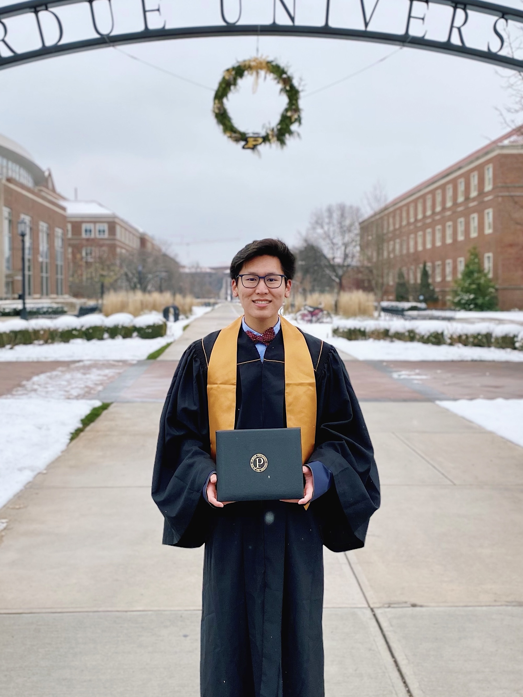 A graduate in Purdue University academic regalia stands on a snowy campus pathway, wearing a black graduation gown with gold stole and holding a diploma. They are wearing glasses and smiling at the camera. Red brick university buildings and winter landscaping are visible in the background. The image is overlaid with text reading 'Purdue University Fall 2020'