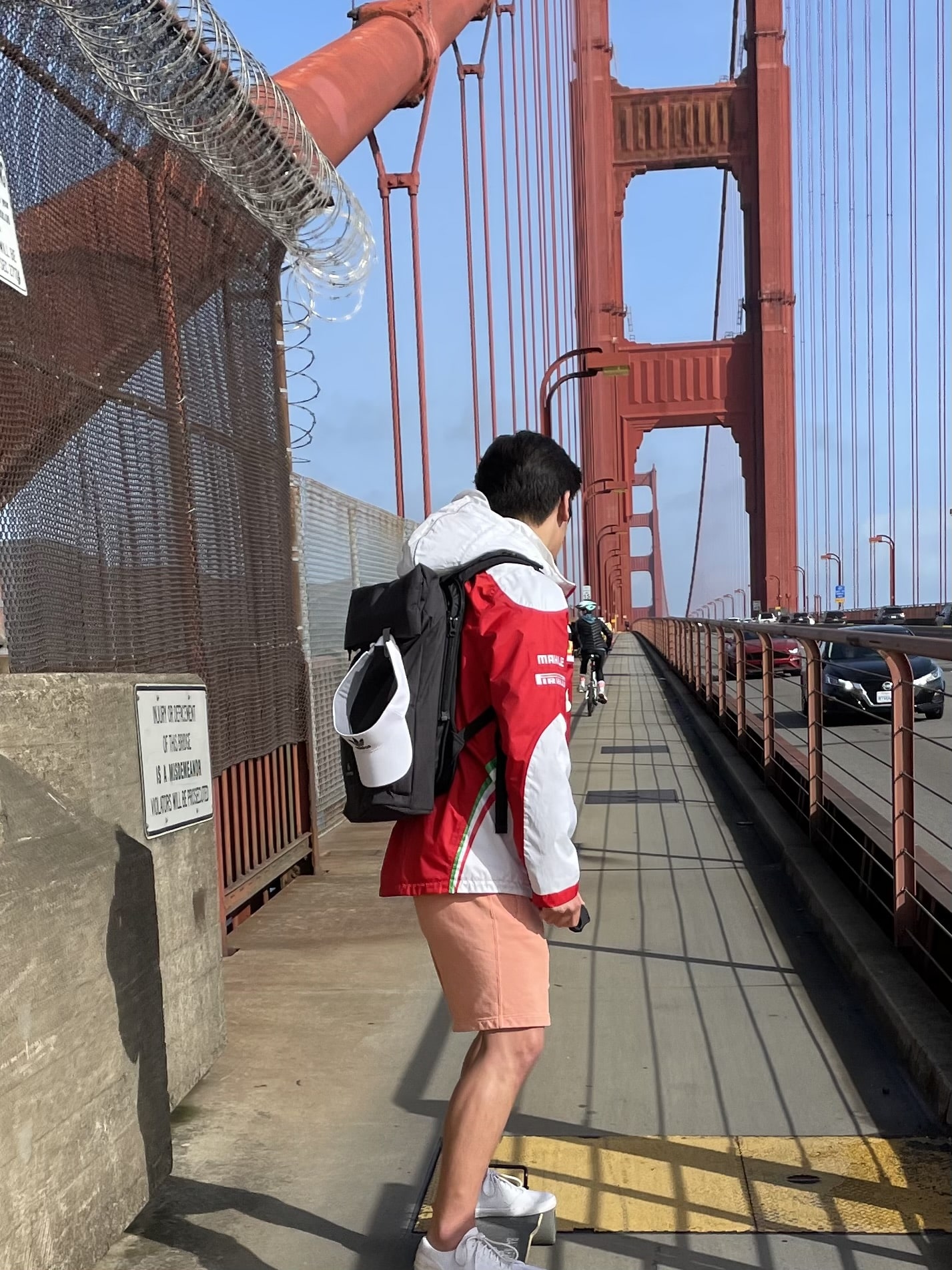 A person standing on an electric longboard on the pedestrian walkway of the Golden Gate Bridge, wearing a red and white jacket, salmon-colored shorts, and carrying a backpack. The iconic red bridge structure and safety railings are visible. The image is overlaid with text reading 'Electric longboard daredevil'