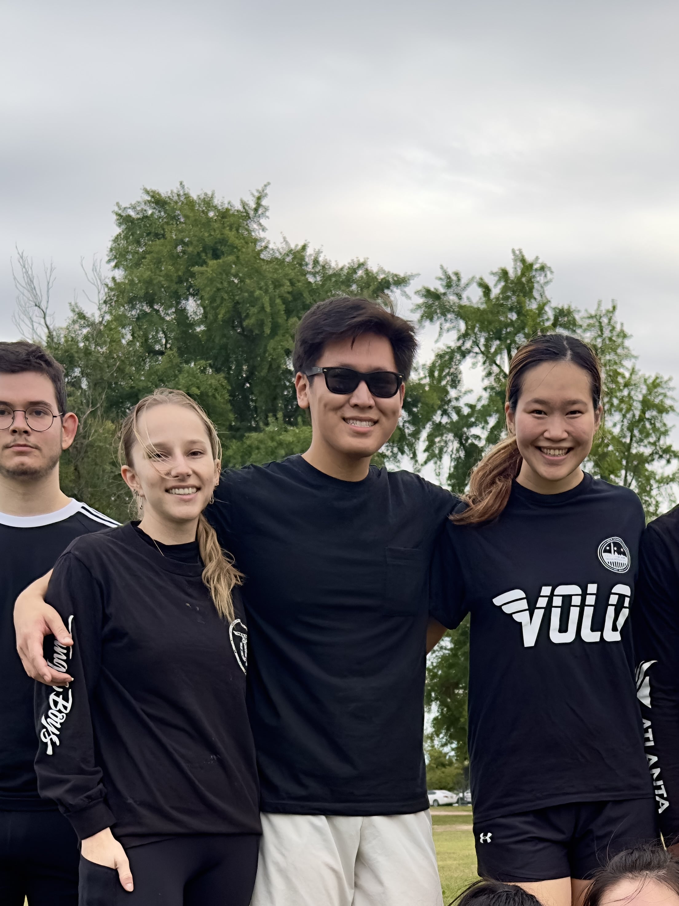 Four people in soccer team attire standing together outdoors. Team members are wearing black shirts with various logos including 'VOLO'. The person in the center is wearing sunglasses and white shorts. Trees are visible in the background on a cloudy day. The image is overlaid with text reading 'Recreational soccer co-captain'