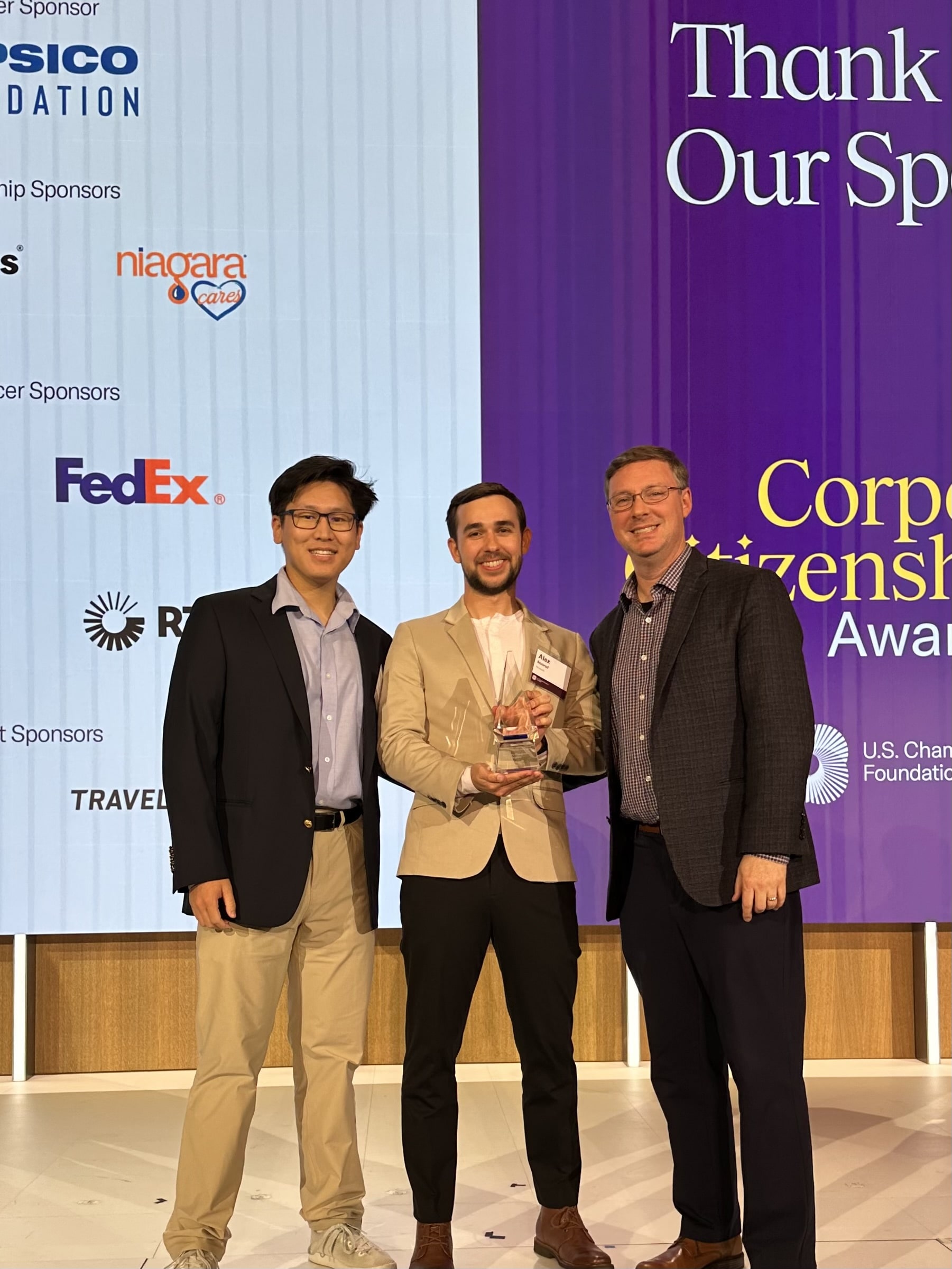 Three people standing on stage at a corporate citizenship awards ceremony. FedEx logos and US Chamber Foundation banners visible in background. The middle person is holding an award trophy, flanked by two others in business attire. All are wearing business casual to formal attire including blazers. The image is overlaid with text reading 'Civic engagement volunteer'